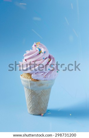 Cupcake in an ice-cream cone - perfect summer dessert for an outdoor birthday party. Strawberry buttercream icing contrasting with sky blue background. Blurry lines from colorful sprinkles.