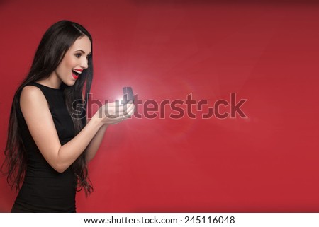 Surprised beautiful woman holding jewelry box with an engagement ring in it. Happy young woman after marriage proposal. Red background.