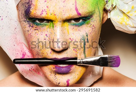 Close-up portrait of young woman with unusual makeup. Model holding makeup brash in her mouth. Woman posing with paint drops over her face. Creative makeup