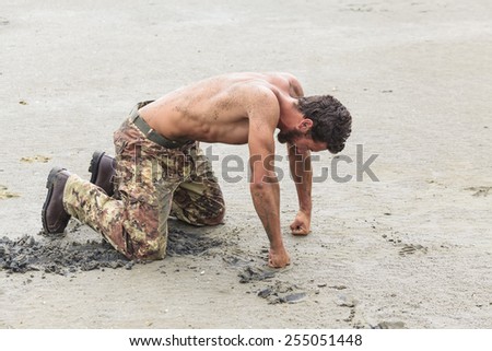 Muscled Shirtless Soldier in Camouflage Pants and Black Shoes Kneeling on the Beach Sand with Fists on the Ground