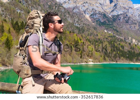 Beard hiker with backpack and camera sitting on railing at the lakeside