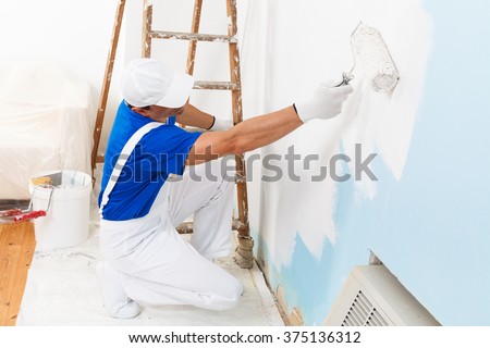 above view of painter with cap and gloves painting a wall with paint roller, wooden vintage ladder and bucket on background