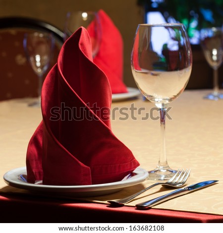 Table in a restaurant with a yellow tablecloth, red napkins, wine glasses and cutlery.