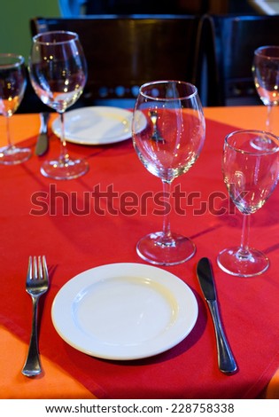 Service for two persons: table in a restaurant with a red and orange tablecloth, wine glasses, white plates and cutlery. (Soft focus.)