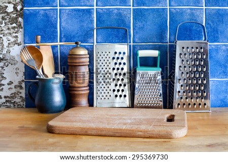 Kitchen utensil. Different kinds stainless steel grater, green jug, vintage wooden spoons, cutting board. Blue tile ceramic background. Wood table.