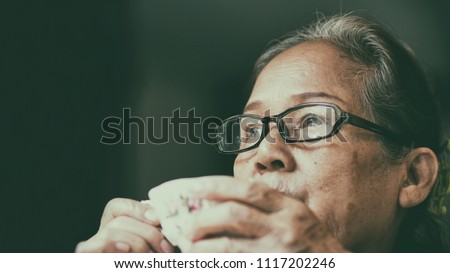 Image of 60s or 70s  Asian elderly drinking tea.Old woman wearing eye glasses,lonely with cold cup.She looking outside or flew away ,Vintage film color tone.Sad elderly concept.