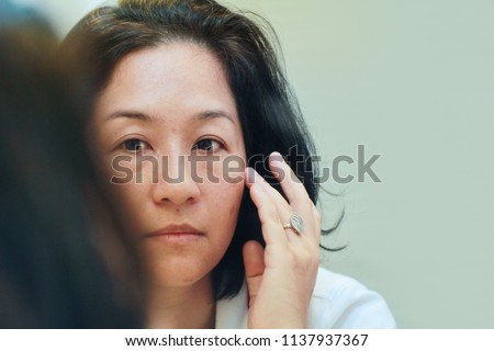 40s Asian woman looking on her face skin in the mirror and look worried or concern about the Aging skin problem.Brownish colored patches or melasma appear on the cheeks.It typically occurs on the face