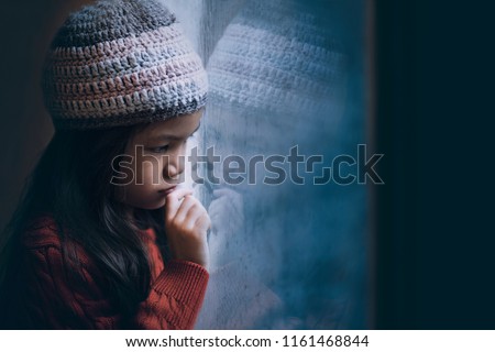 6 years old Asian girl sad by the window in winter red sweater and knitted hat.Cold weather make condensation on window glass,girl draw sad face on it.Concept of sad child.Preteen lifestyle.