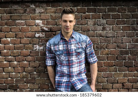 Smiling young man leaning on old brick wall, wearing blue and red shirt and jeans
