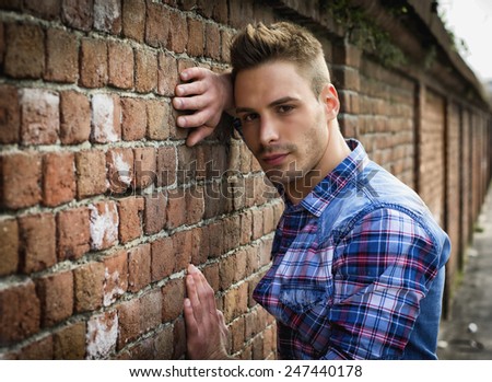 Young man leaning on old brick wall, wearing blue and red shirt