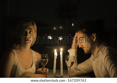 A couple appear to be in a state of tension as if grappling with a serious problem. They are drinking white wine and sit at a table lit by candlelight.
