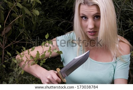 An attractive blonde woman cuts her way through thick bushes with a large knife. The bush is symbolic of life\'s difficulties./Woman with knife/