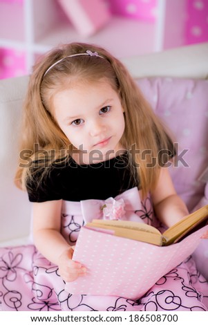Charming little girl wearing beautiful pink dress reading book with her on white sofa in bright pink room