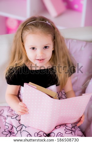 Charming little kid wearing beautiful pink dress reading book with her on white sofa in bright pink room