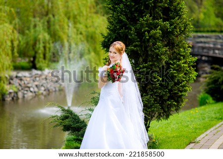 Pretty bride in a park on the background of the fountain with a red wedding bouquet in hands