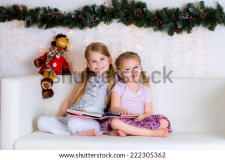 two sisters happy with a new book of Christmas decorations near a Christmas tree on a white sofa at home