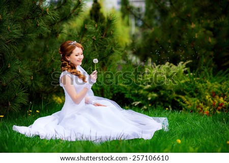 beautiful bride in a pine forest sitting on the grass and looking at dandelion, bee flying over flower