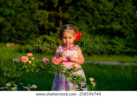 little girl in a park next to a bush of roses with red bows looking at flowers