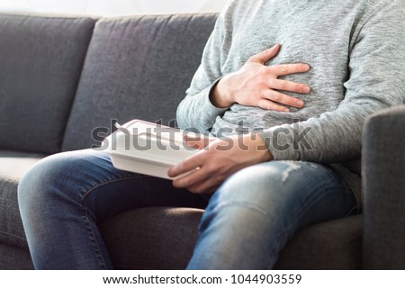 Stomach pain, food poisoning or digestion problem after fast junk food. Man ate too much and is holding belly with hand. Indigestion, heart burn or unhealthy diet.
