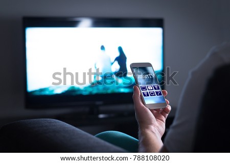 Man watching television and using smart tv remote control application on mobile phone. Choosing movie stream, switching channel or changing settings in the menu and user interface with smartphone.