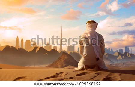 Arab Emirati man praying on top of a dune in UAE desert front Dubai skyline.