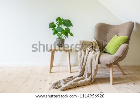 Beige armchair with a green pillow, blanket and a wooden table with a potted plant, fruit salad tree (Monstera deliciosa). Empty white wall in simple living room interior. Copy space