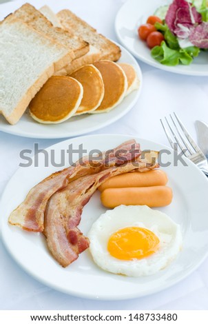 set of American breakfast including fried egg, sausage and bacon serves with salad, bread and pancakes on white table