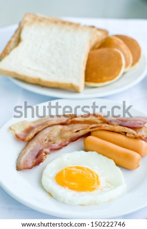 set of American breakfast including fried egg, sausage and bacon serves with bread and pancakes on white table