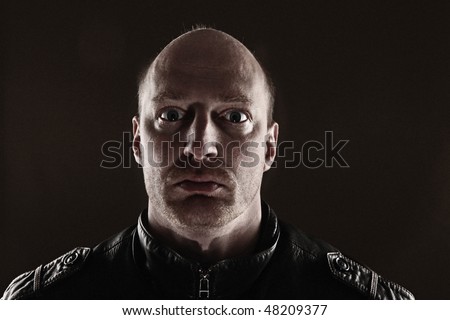 portrait of male skinhead in leather jacket lit from behind. Dark picture of bald man with stubble