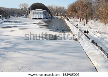 Winter snow-covered park. Partially frozen pond. Floating birds. The building of a summer theater