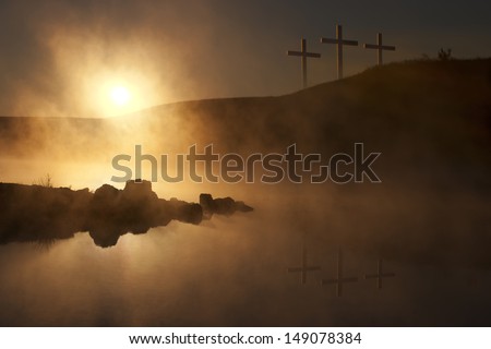 Dramatic religious photo illustration of Easter Sunday Morning reflecting a prayerful moment as a warm sun rises over a foggy lake, and three crosses on a hill reflect in the water below.