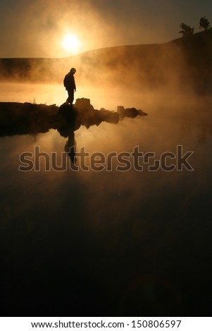 A warm, foggy sunrise silhouettes a hiker against a cold early morning mist across a lake.
