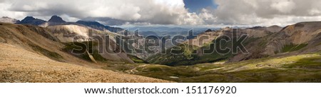 Panoramic view of a Rocky Mountain pass in Colorado includes high altitude meadow, tree lined ridges, fields of weathered stones, crooked jeep trails, billowing clouds, and distant peaks and valleys.