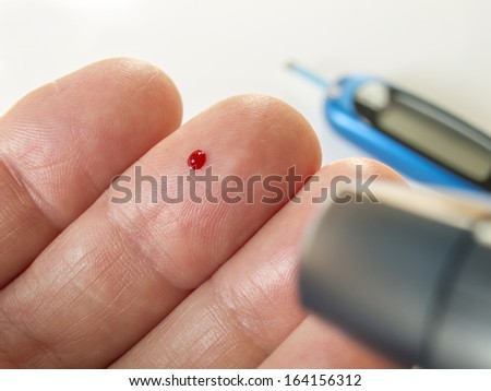 Photo of someone using a Type II Blood Sugar Testing Finger prick device. Out of focus in the background is the meter with testing strip to give the results.