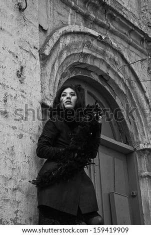 Mysterious young woman in black coat and fur standing next to  old building