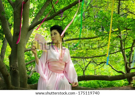 Japanese young beautiful woman wearing  pink kimono with fan sitting on a tree branch and looking into the distance with playful smile in blooming garden. Spring green forest. Colorful silk ribbons