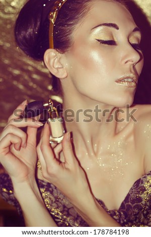 Portrait of young beautiful woman with gold makeup holding perfume spray on the gold background. Brunette with retro hairstyle and hoop. Shiny skin texture. Luxury