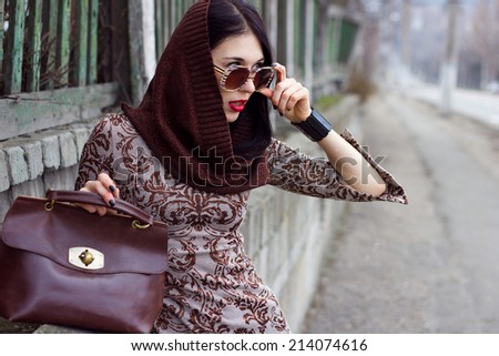 Young beautiful woman in warm clothes and glasses with  brown leather retro bag looking into the distance posing outdoors. Season Autumn Winter . Fashion model shooting.