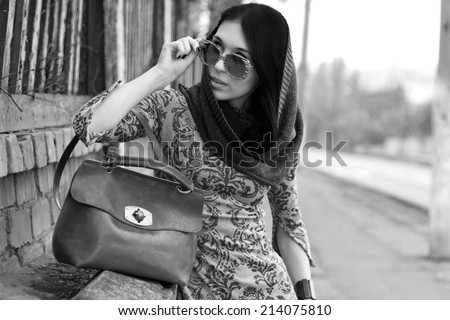 Young beautiful woman in warm clothes and glasses with brown leather retro bag looking into the distance posing outdoors. Season Autumn Winter 2014. Fashion model shooting. Black and white image.