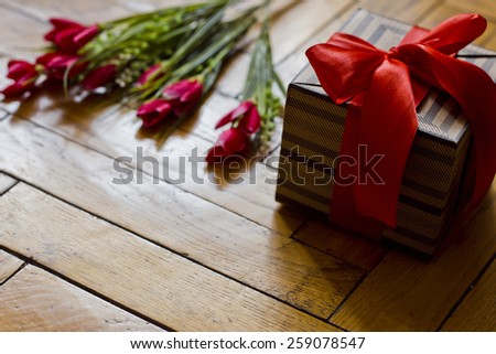 Open gift box present with red bow and beige brown strips on wooden background. Bouquet of red scarlet flowers tulips. Romantic style. Holidays.