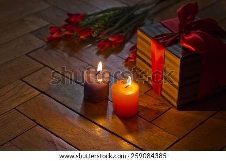 Burning candles, open gift box present with red bow and beige brown strips, bouquet of red scarlet flowers tulips on wooden background. Romantic style. Holidays.