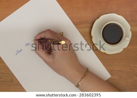 Female hand writing letter on white office paper on wooden desk.
