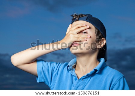 Teenage boy, standing outside, its evening, his hand covering his eyes, squinting out of his left eye.