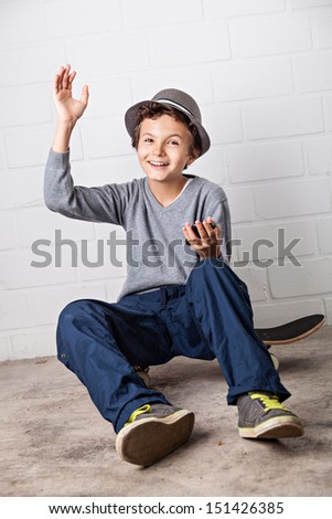 A Teenage boy, sitting on his skateboard, holding a smartphone in his hands with which he is playing around with. He is waving with his right hand