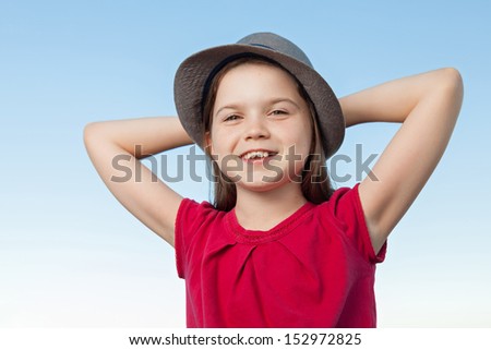 A portrait of a cute little girl, she is standing outside, wearing a hat and a red shirt against a blue sky, her arms are crossed behind her head, she looks relaxed and very happy