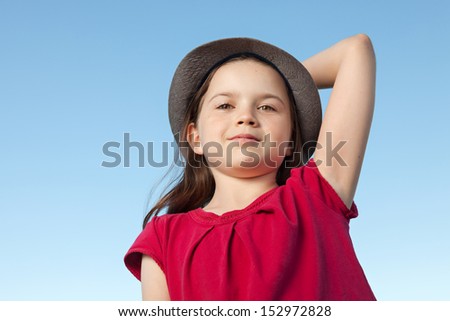 A portrait of a cute little girl, she is standing outside, wearing a hat and a red shirt against a blue sky,