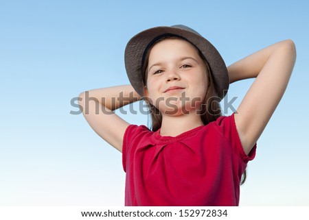 A portrait of a cute little girl, she is standing outside, wearing a hat and a red shirt against a blue sky, her arms are crossed behind her head, she looks relaxed and confident