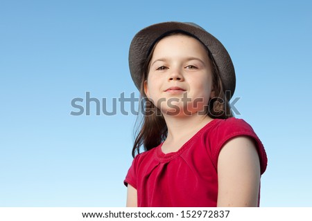 A portrait of a cute little girl, she is standing outside, wearing a hat and a red shirt against a blue sky,