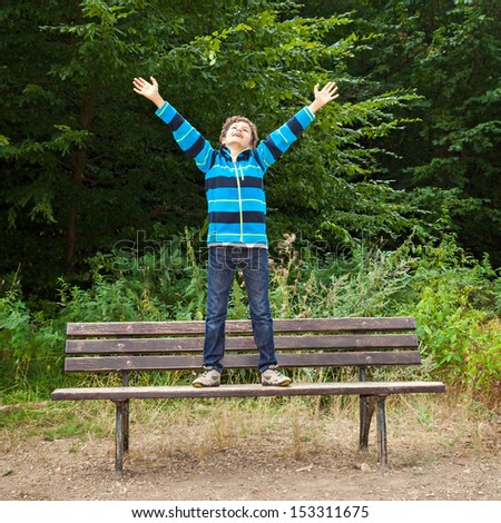 A teenage boy in a forest is standing on a bench, his arms up in the air, he looks very happy.