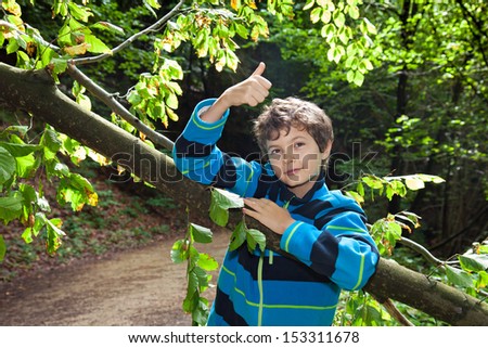 A teenage boy in a forest is very happy, smiling very friendly and giving the thumbs-up sign with his right hand.